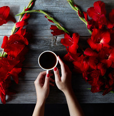Cup of hot black coffee in female hands on a wooden vintage desk with summer red flowers gladioli. The concept of coffee on a summer day.