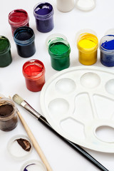 Open gouache jars with empty white plastic palette and few paintbrushes on a white background,...