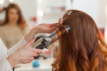 Selbstklebende Fototapeten Friseursalon Hairdresser makes hairstyle girl with long red hair in a beauty salon. Create curls with curling irons. Professional hair care.  © Dimid