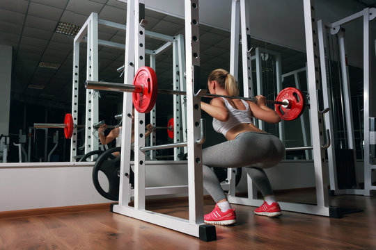 Young sexy blonde european girl in the gym doing squat with barbell in front of the mirror. She is dressed in sports clothes white top, gray leggings, orange sneakers 
