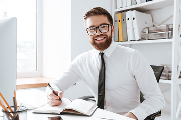 Cheerful businessman sitting in office while writing notes