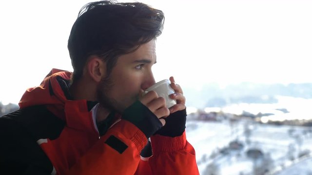 Young man sipping from a cup of hot coffee on a wooden deck in winter and admiring landscape