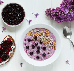 Healthy breakfast - oatmeal porridge with berries, nuts and seeds and toast with currant jam on a white wooden table with spring flowers. Useful vitamins nutrition.