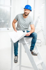 Portrait of a handsome builder, foreman or repairman in the helmet sitting with drawings on ladder...