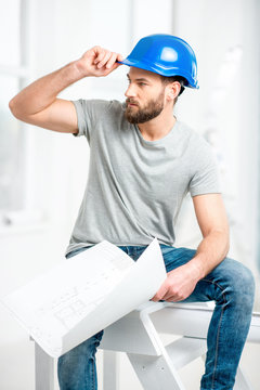 Portrait of a handsome builder, foreman or repairman in the helmet sitting with drawings on ladder in the white interior