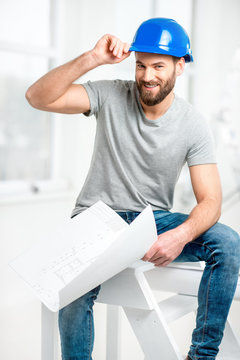 Portrait of a handsome builder, foreman or repairman in the helmet sitting with drawings on ladder in the white interior