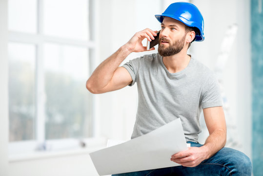 Handsome repairman or builder in helmet talking with phone with drawings in the white interior