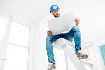 Portrait of a handsome builder, foreman or repairman in the helmet sitting with drawings on ladder...