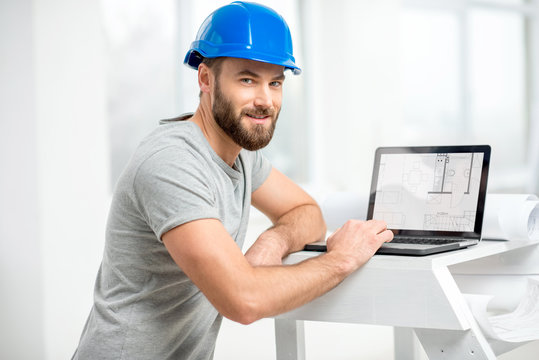 Handsome architect or foreman in helmet working with laptop on the apartment drawings at the structure white interior