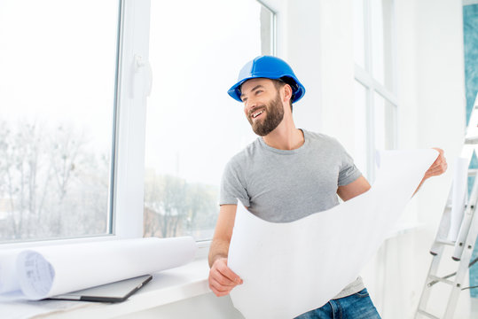 Portrait of a handsome builder, foreman or repairman in the helmet looking at the paper drawings near the window indoors