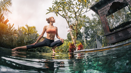 Young woman performs yoga exersises in the tropical garden by the pool