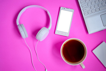 Overhead of modern comfort work place. Different objects on pink background. Items include laptop, earphones, smartphone and cup of coffee.