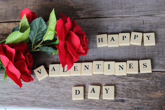 Happy Valentines day wording by crossword on old wooden board with artificial red roses background.