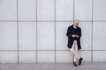 Fototapeta na wymiar Young handsome bald businessman holding a smart phone, looking down tapping screen leaning on a white wall - technology, business, work concept 