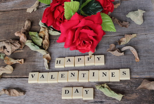 Happy Valentines day wording by crossword on old wooden board with artificial red roses background. Love and romance concept.