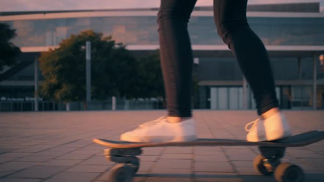 Camera follows girl's legs in tight black jeans skating on small surfskate longboard on urban street at sunset from left to right