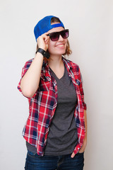 Hipster girl with short hair in a red plaid shirt, gray t-shirt, blue jeans, dark sunglasses, a blue cap, a black bracelet on a white background smiling and holding his hand over his glasses. 