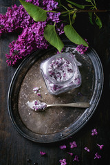 Obraz na płótnie Canvas Arrangement with glass jar of lilac flowers in sugar, silver spoon and lilac branch on vintage tray over black wooden background. Flat lay. Dark rustic atmosphere.