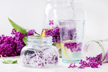Obraz na płótnie Canvas Glass jar of lilac flowers in sugar, glass and pitcher of lilac water with lemon and branch of fresh lilac on white linen tablecloth.