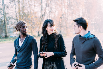 Three multiethnic woman and men friends walking outdoor in city back light, chatting and holding smart phone - friendship, social network, conversation concept