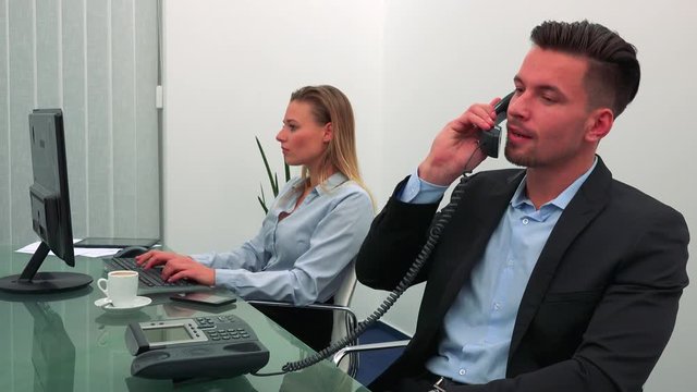 A young, handsome office worker answers the phone, his colleague works on a computer in the background