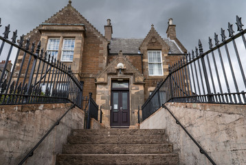 Orkneys, Scotland - June 5, 2012: The facade of Stromness town hall whick stands up a flight of stairs. Brown and gray building under dark cloudy skies. Black fence and gray stone stairs.