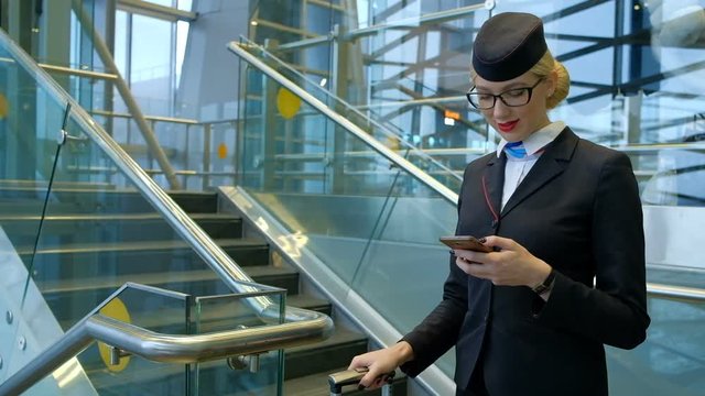Stewardess at the airport with a phone in her hands helps passenger. Worker in blue uniform with one hand writes a sms on your smartphone near the stairs, then it diverts young tall blonde in glasses