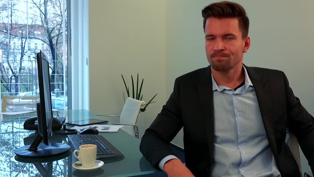 A young, handsome man sits at a desk in an office, looks at the camera and shakes his head