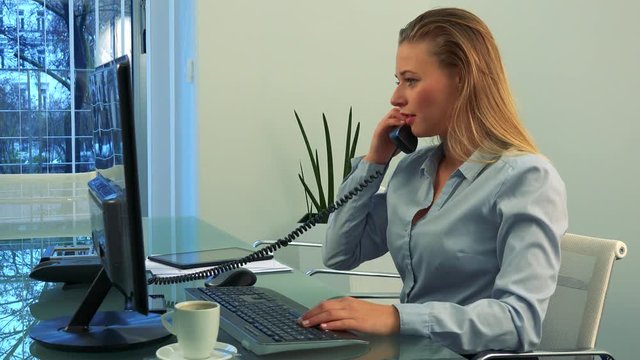 A young, beautiful woman works on a computer in an office, then picks up the phone receiver and talks