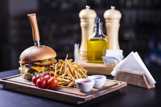 Tasty grilled cheeseburger with a knife stabbed trough on a wooden plate. Burger with pickles, cheese, tomatoes and french fries on the side. Napkins and olive oil in the background.