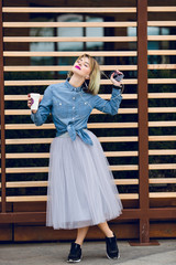A standing blonde girl with pink lips holding a cup of coffee and listening to music on a smartphone with striped wooden balks behind wearing blue jeans shirt, grey tulle skirt and black sneakers