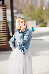 Fototapeta premium A portrait of a standing dreamy blonde girl with bright pink lips hoding her hand near chin wearing denim shirt, grey tulle skirt and black watch