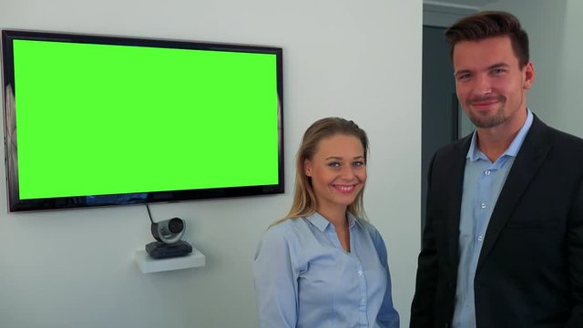 A man and a woman (both young and attractive) stand beside a green television screen and smile at the camera