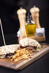 Tasty grilled sandwiches with a wooden stick stabbed trough them on a wooden plate. Meat sandwiches with lettuce, tomatoes and french fries on the side. Napkins and olive oil on the background.