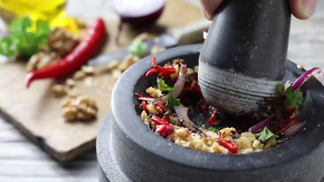Assorted fresh herbs, nuts and spices in a mortar and pestle being ground for use as condiments in cooking a savory recipe in a close up view with ingredients behind on a chopping board