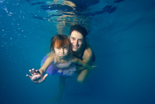 The little girl and the instructor swim underwater, smiling and looking at camera against a blue background. Portrait. Close-up. The view from under the water. Landscape orientation