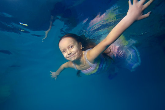 Little girl in a beautiful dress swims underwater, arms outstretched, looking at camera and smiling. Portrait. Close-up. Shooting under the water surface. Horizontal view
