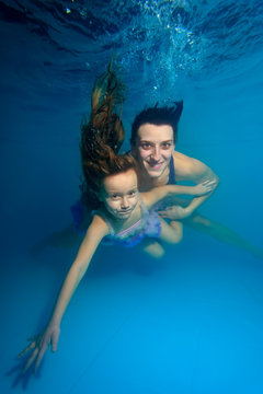 Mother and daughter with disheveled hair floating underwater on a blue background, dive to the bottom of the pool, looking at the camera and smiling. Portrait. Vertical view