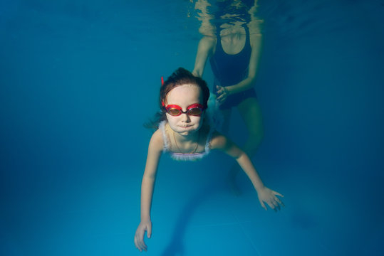 The child, a little girl is engaged in and swims underwater in the pool, and the coach helps her. Portrait. Shooting under the water surface. Horizontal view