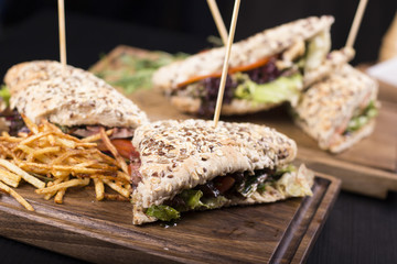 Delicious sandwiches, tomatoes, and french fries. Pickles and lettuce dripping off the side. Barbecue stick stabbed through the sandwiches on a wooden table.