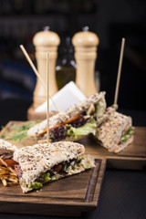 Tasty grilled sandwiches with a wooden stick stabbed trough them on a wooden plate. Meat sandwiches with lettuce, tomatoes and french fries on the side. Napkins and olive oil on the background.