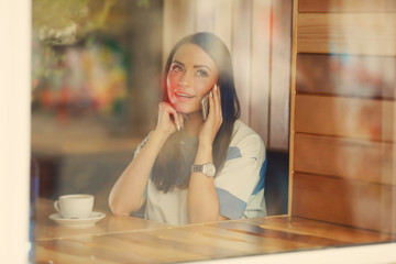 Beautiful young woman talking by mobile phone while sitting in cafe, view from outside