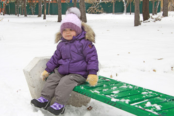 Child in lilac winter clothes sitting on a green bench in snow