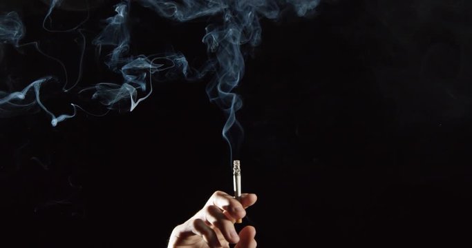 Elegant smoke rises from cigarette in hand, slow motion against black background