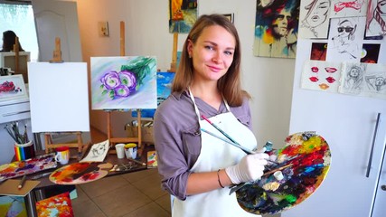 Pretty Young Teacher of Art School Smiling to Camera With Brushes in Hand, Which Holding in Palette in Art Studio Hung With Sketches and Drawings.