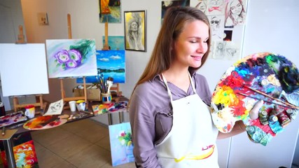 Women Beautiful Painter Posing and Smiling Into Camera With Brushes and Palette in Art Studio Hung With Sketches and Drawings.