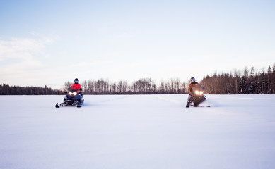 A father and his teenage daughter wearing helmets and winter gear racing side by side on snowmobiles across a white snow covered field with forest of trees in the background in a winter landscape