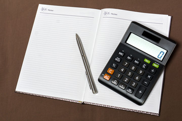 Notebook, calculator  and silver pen on brown cloth background.