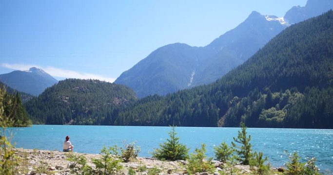 Woman Sitting & Relaxing in Solitude by Clear Blue Lake surrounded by Mountains
