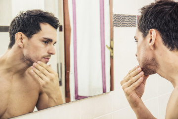 Shirtless handsome young man examining his stubble in bathroom mirror in the morning